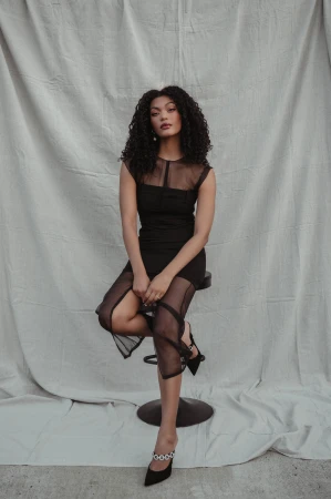 Woman with curly dark hair wearing a black sheer panel dress seated on a stool against a textured white fabric backdrop