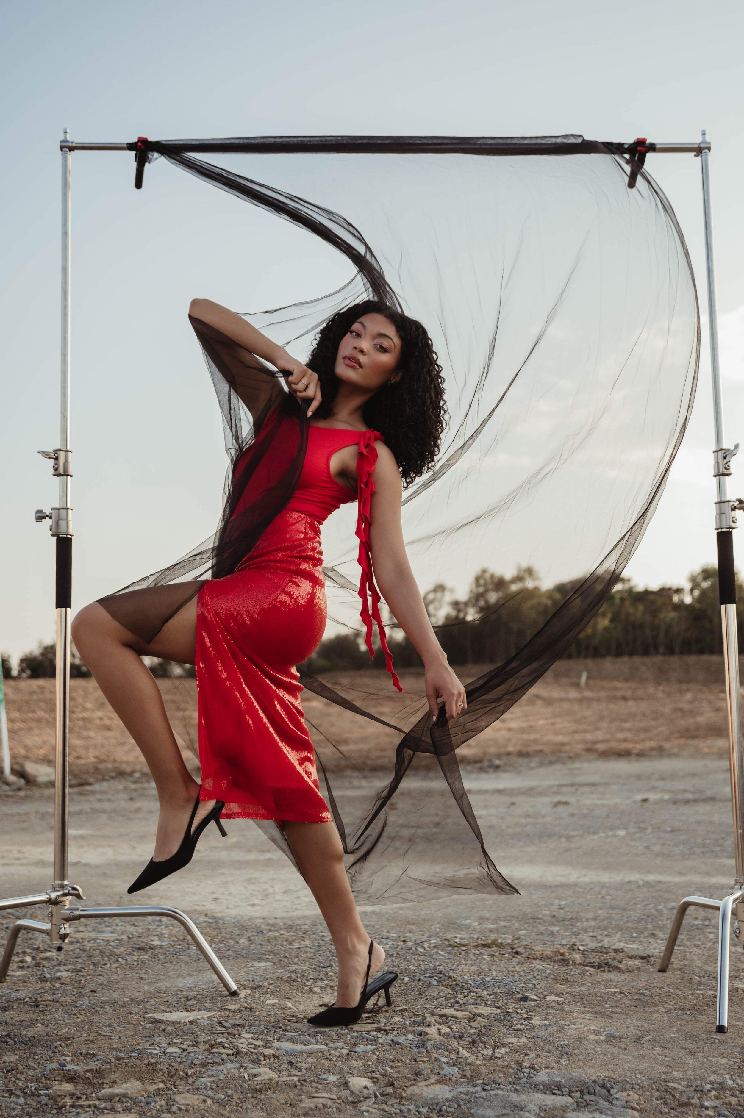 A woman poses in a red dress with black tulle in an outdoor shot with a blue sky, wearing black high heels and standing on gravel.