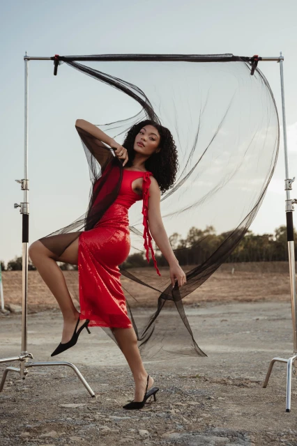 A woman poses in a red dress with black tulle in an outdoor shot with a blue sky, wearing black high heels and standing on gravel.
