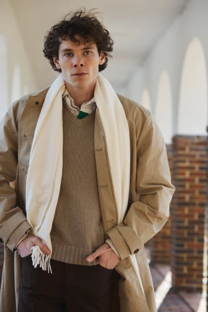 Man in beige trench coat and sweater, wearing a cream-colored scarf, poses in a covered walkway with arched columns and brick pillars.