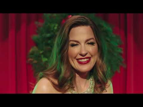 Smiling adult woman with red lipstick and necklace poses in front of a green leafy background and red curtains, eye-level portrait.
