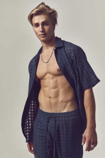 Athletic blond male model poses shirtless in an open dark-blue patterned button-down shirt and matching pants in a studio setting.