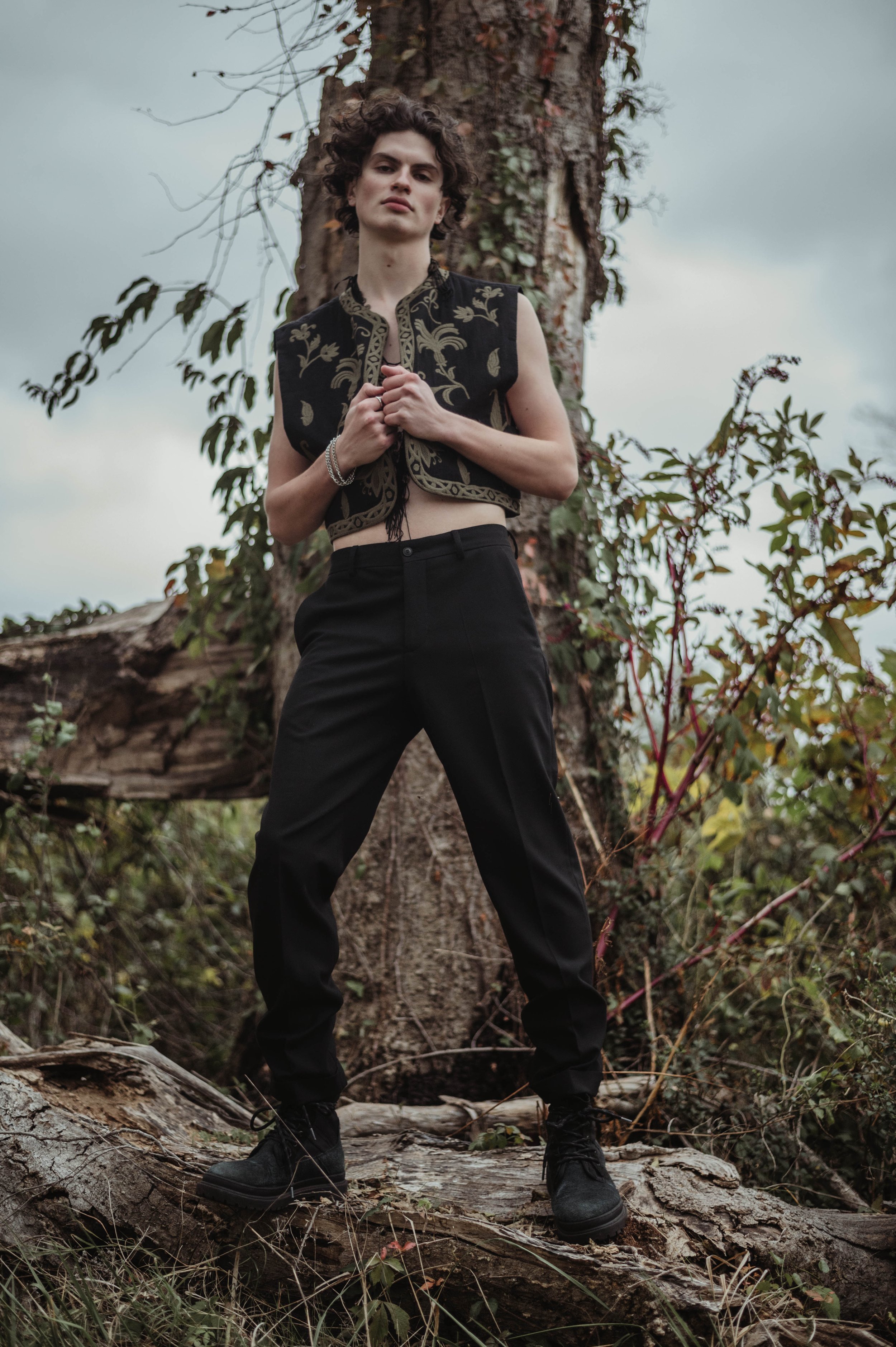 Male fashion model poses in embroidered vest and black pants near a fallen tree in an outdoor setting with an overcast sky.