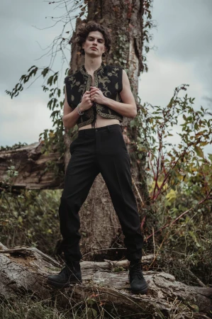 A young man with curly dark hair posing outdoors in a black embroidered vest and black trousers standing in front of a large tree.