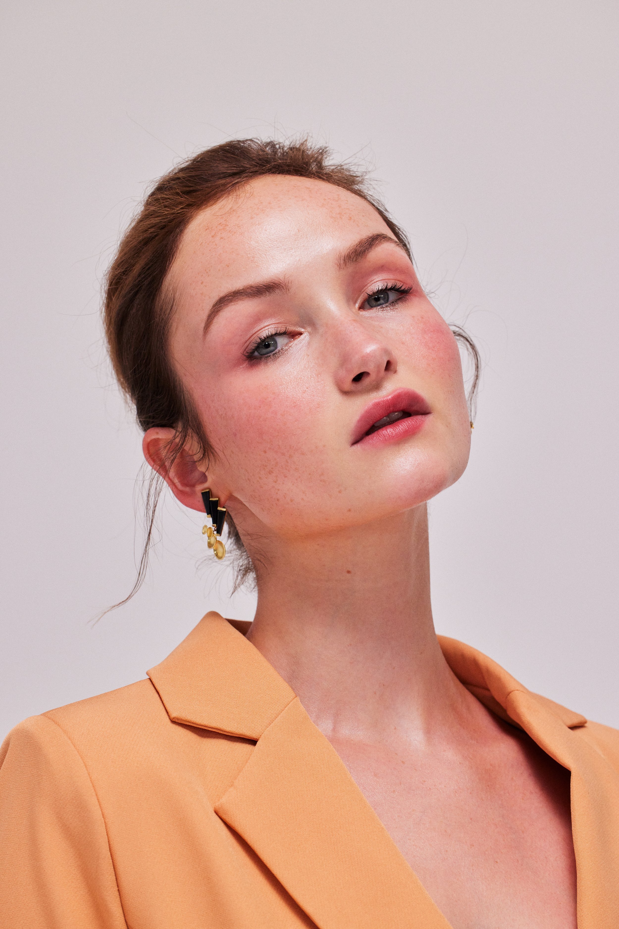 Portrait of a red-haired woman with fair skin in a peach blazer, wearing earrings and makeup, against a plain light background.