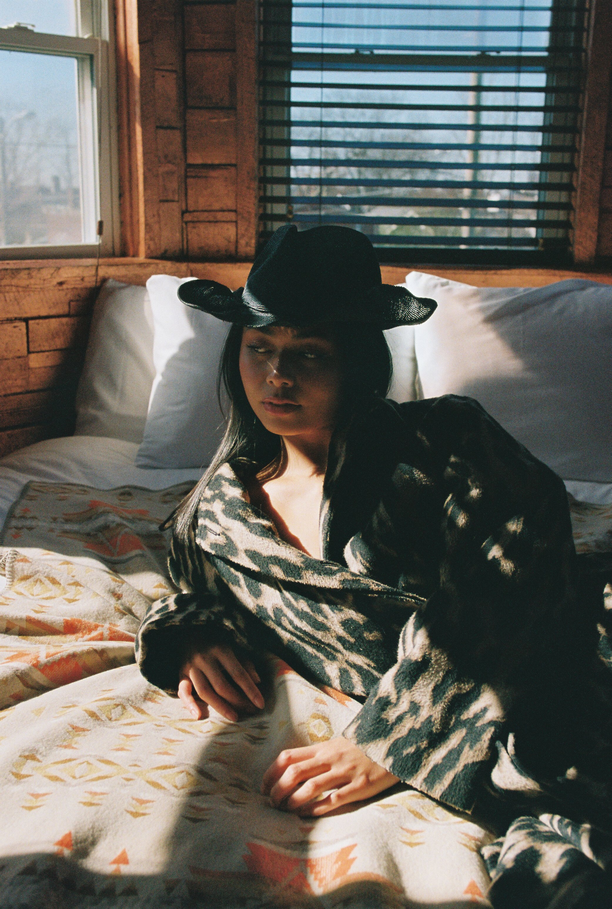 Young woman in animal-print jacket and hat lounges on patterned bed in warm light, looking off to the side in a relaxed pose.