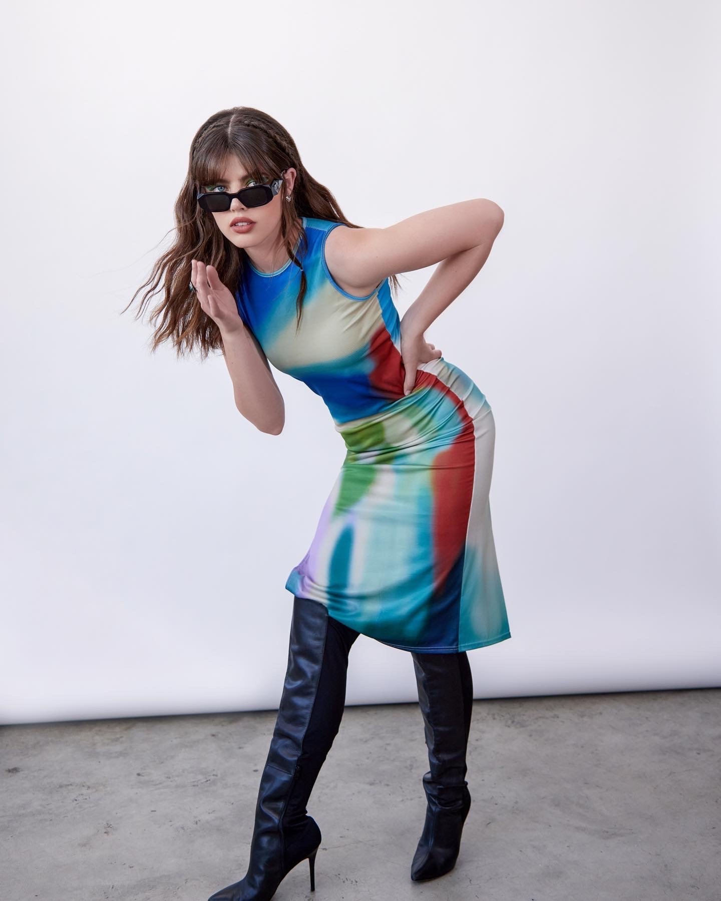 Adult woman poses in studio wearing colorful dress, black thigh-high stiletto boots and sunglasses, against white backdrop.