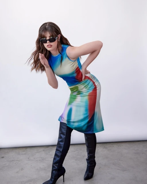 Adult woman poses in studio wearing colorful dress, black thigh-high stiletto boots and sunglasses, against white backdrop.