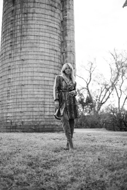 Monochrome portrait of a woman in a dress and boots, posing in front of a tall concrete silo on a grassy field with trees.