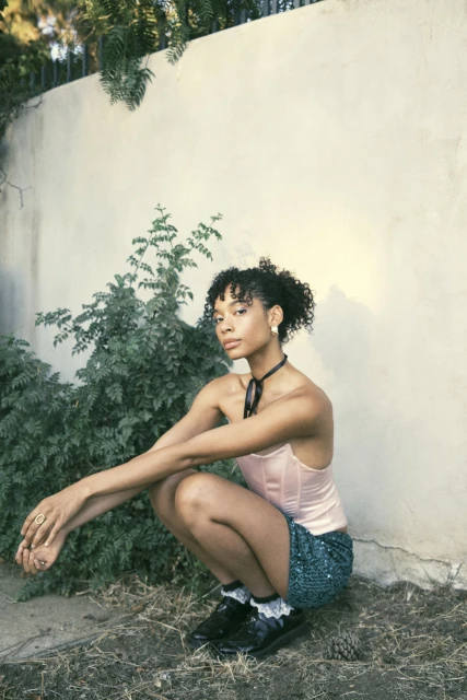 An adult female with curly dark hair poses in a garden squatting beside a stucco wall and plants. She wears a shiny skirt.