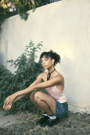 An adult female with curly dark hair poses in a garden squatting beside a stucco wall and plants. She wears a shiny skirt.