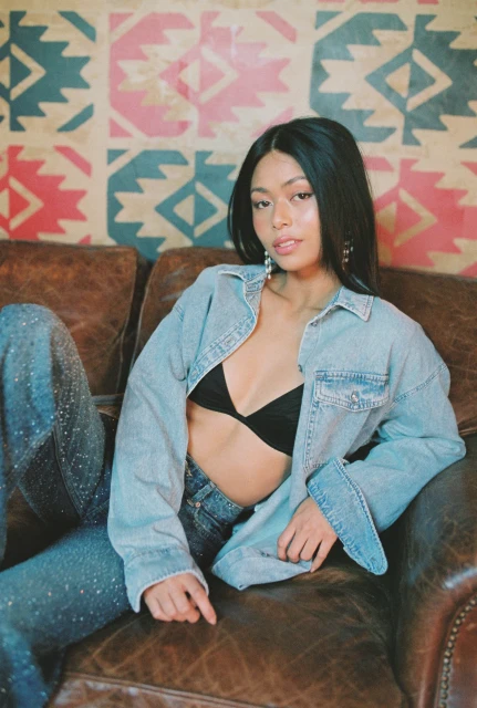 Young woman with black hair poses on a brown leather couch wearing an unbuttoned denim jacket and jeans in an indoor setting.