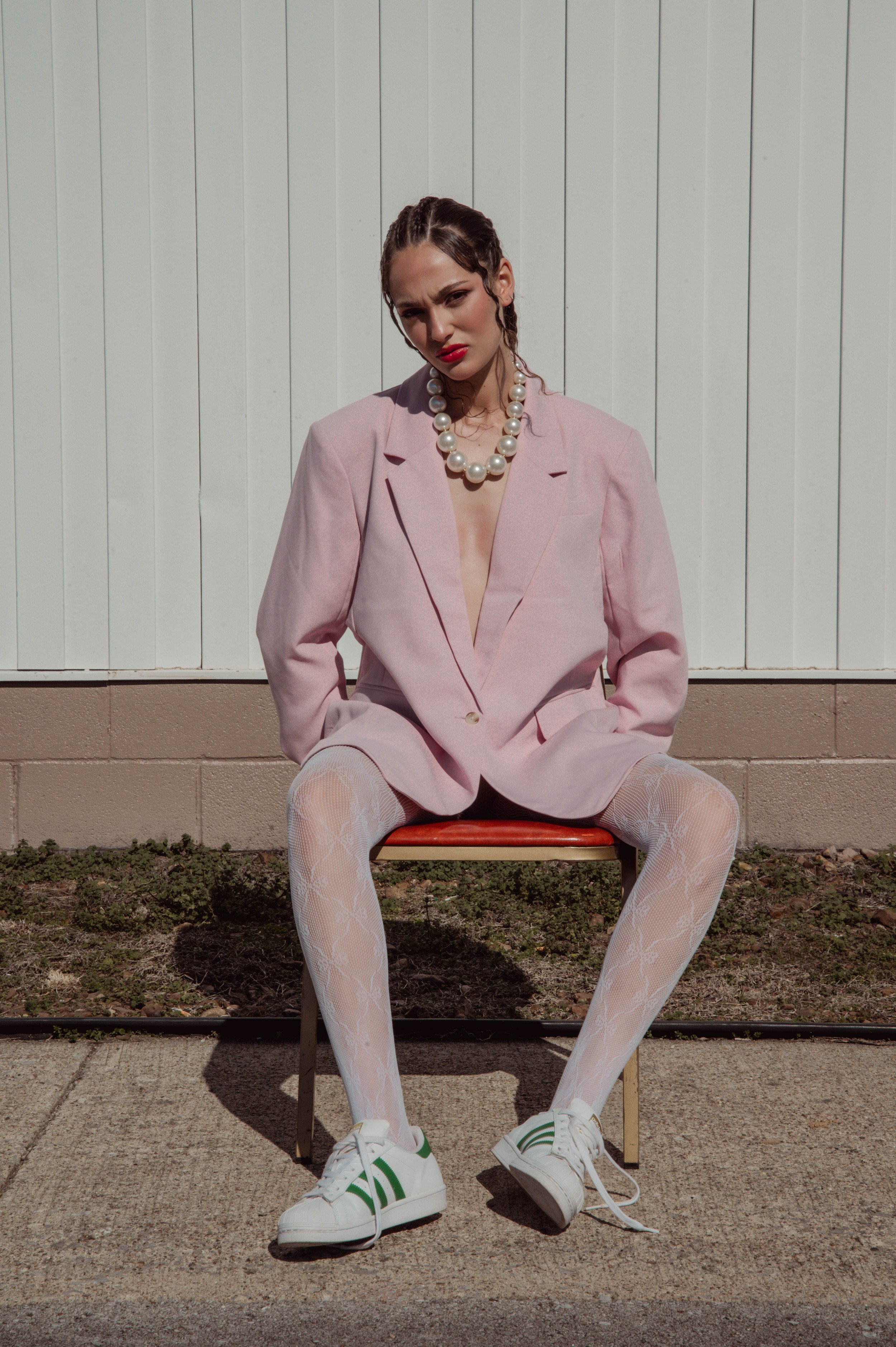 Woman in oversized pink blazer wearing white patterned tights and sneakers seated on a red chair against white vertical panels.