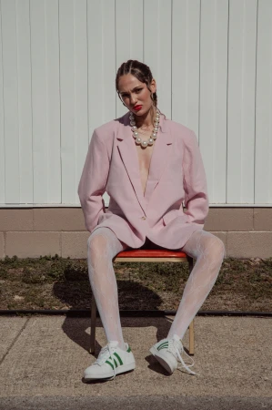 Woman in oversized pink blazer wearing white patterned tights and sneakers seated on a red chair against white vertical panels.
