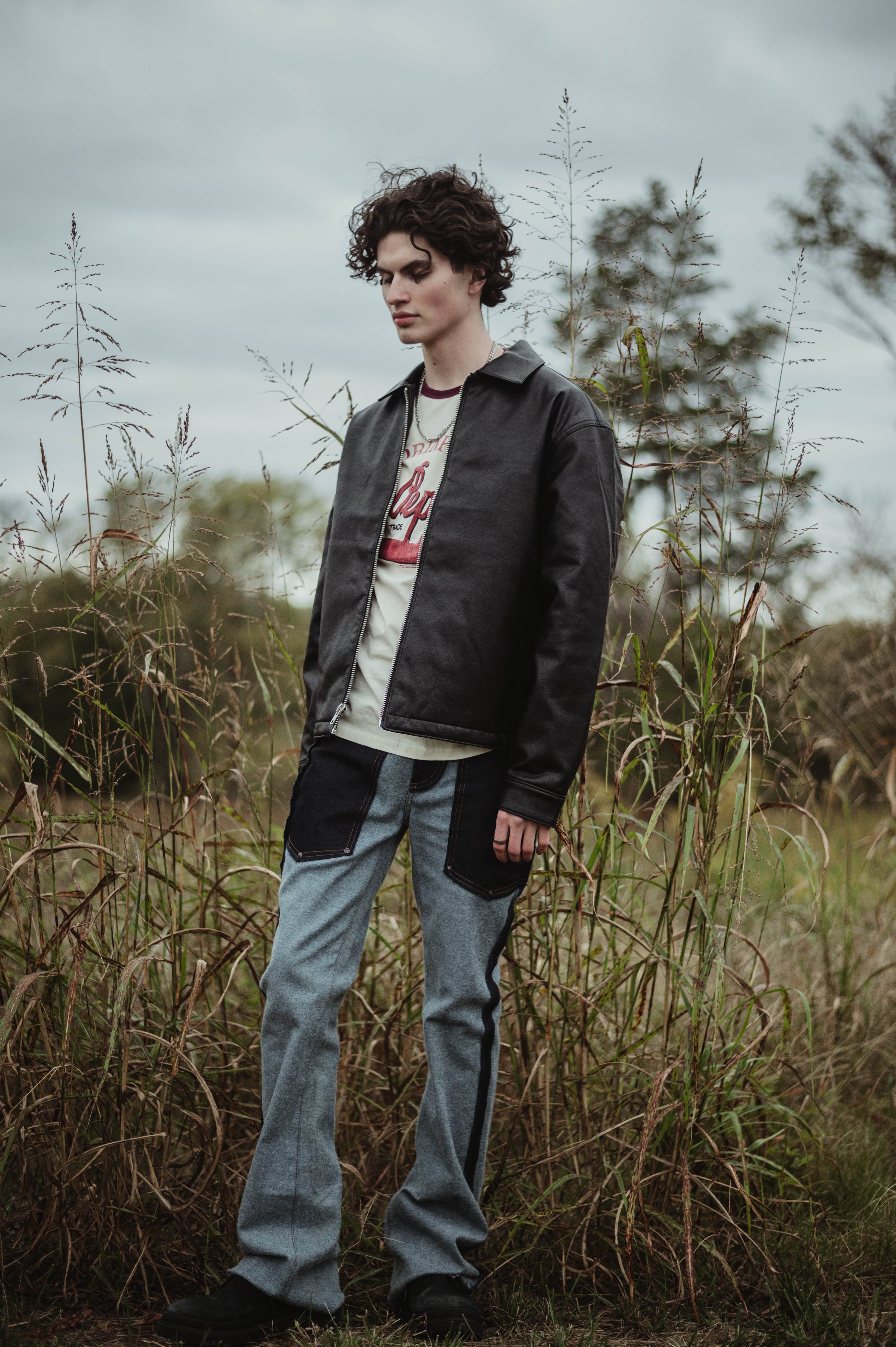 Fashionable adult male with dark curly hair wearing a black jacket and jeans stands in a field of tall grass under an overcast sky.