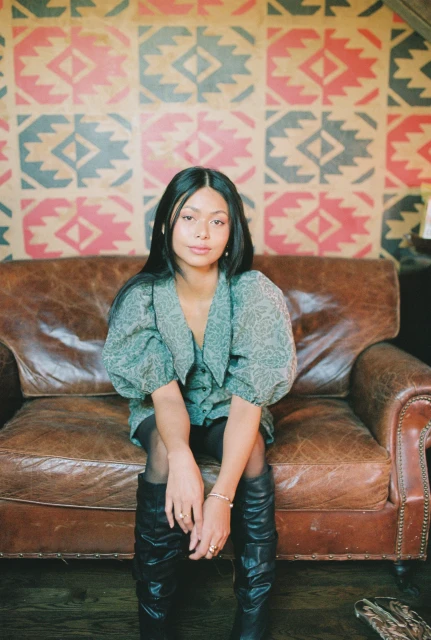 A young adult woman poses on a leather couch in front of patterned wallpaper, wearing a patterned top and black boots in an indoor setting.