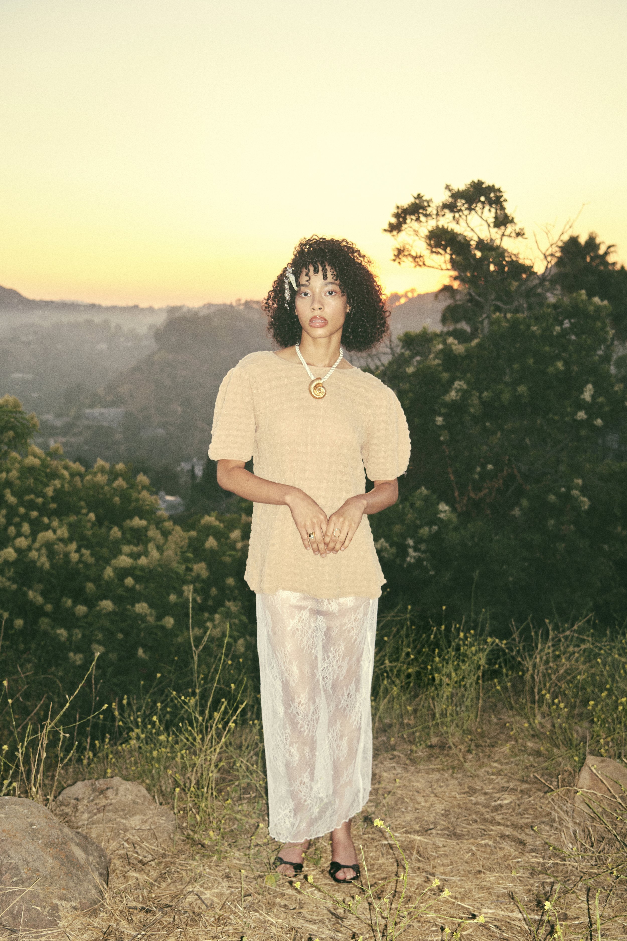 Woman with curly hair wearing a textured beige top and a white lace skirt standing outdoors at sunset.