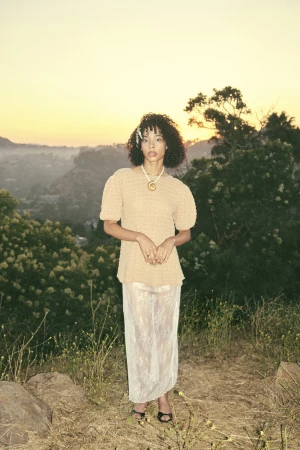Woman with curly hair wearing a textured beige top and a white lace skirt standing outdoors at sunset.