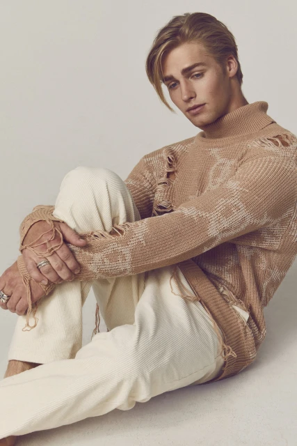 Portrait of a blond adult male in a distressed sweater and pants, sitting in a photo studio with a neutral backdrop, posing for a fashion shot.