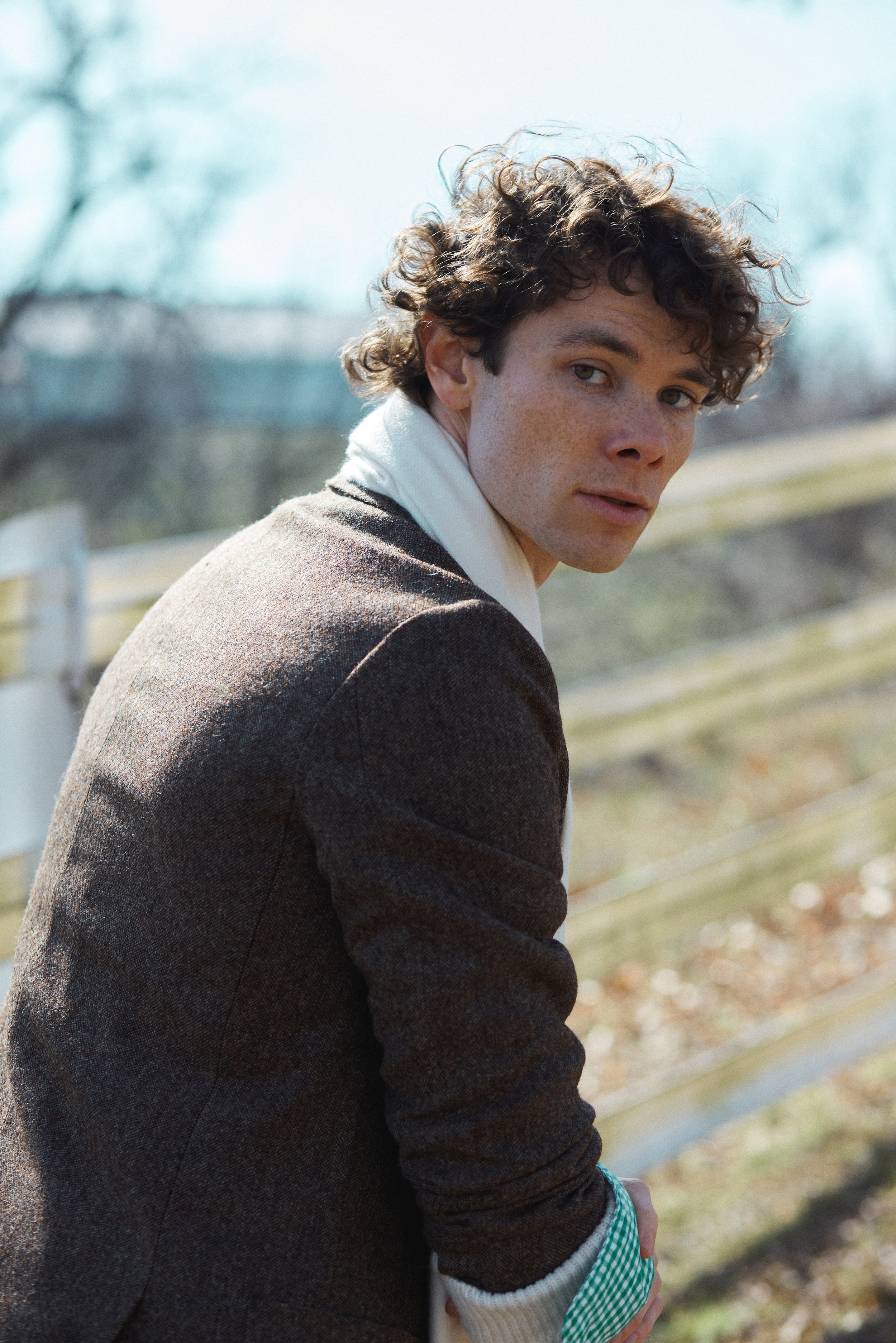 Portrait of a handsome adult male with curly brown hair wearing a brown coat and white scarf, looking back over his shoulder outdoors.