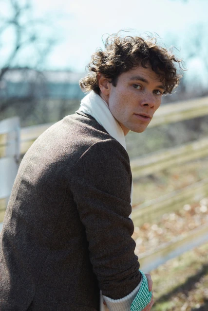 Portrait of a handsome adult male with curly brown hair wearing a brown coat and white scarf, looking back over his shoulder outdoors.
