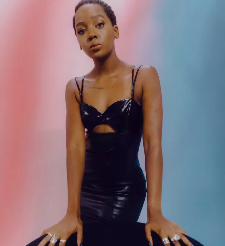 Stylish young woman with dark hair posing in black dress with cut-out, rings on fingers, studio background featuring pink and blue hues.
