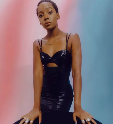 Stylish young woman with dark hair posing in black dress with cut-out, rings on fingers, studio background featuring pink and blue hues.