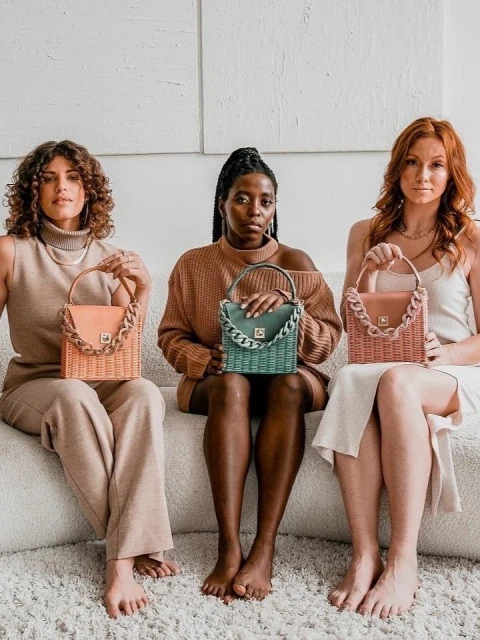 Three women of diverse ethnicities sit together on a cream couch, displaying matching woven handbags with chain-link handles in a light, neutral setting.