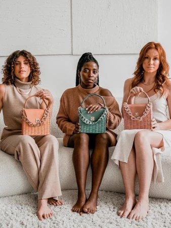 Three women of diverse ethnicities sit together on a cream couch, displaying matching woven handbags with chain-link handles in a light, neutral setting.