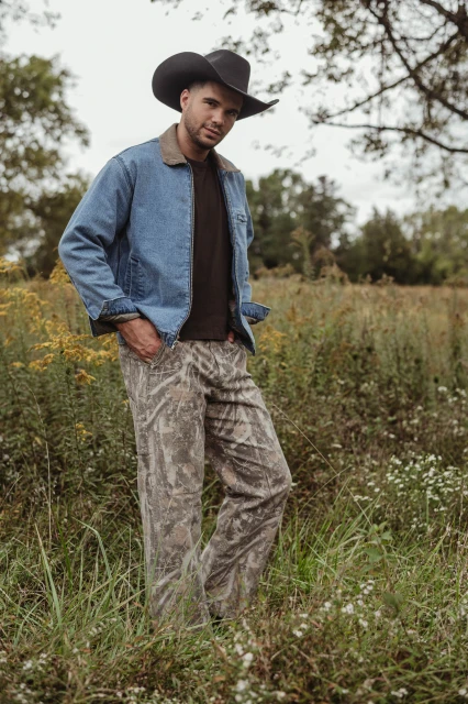 Man in a cowboy hat and denim jacket standing in a field of tall grass with trees in the background, wearing camouflage pants.