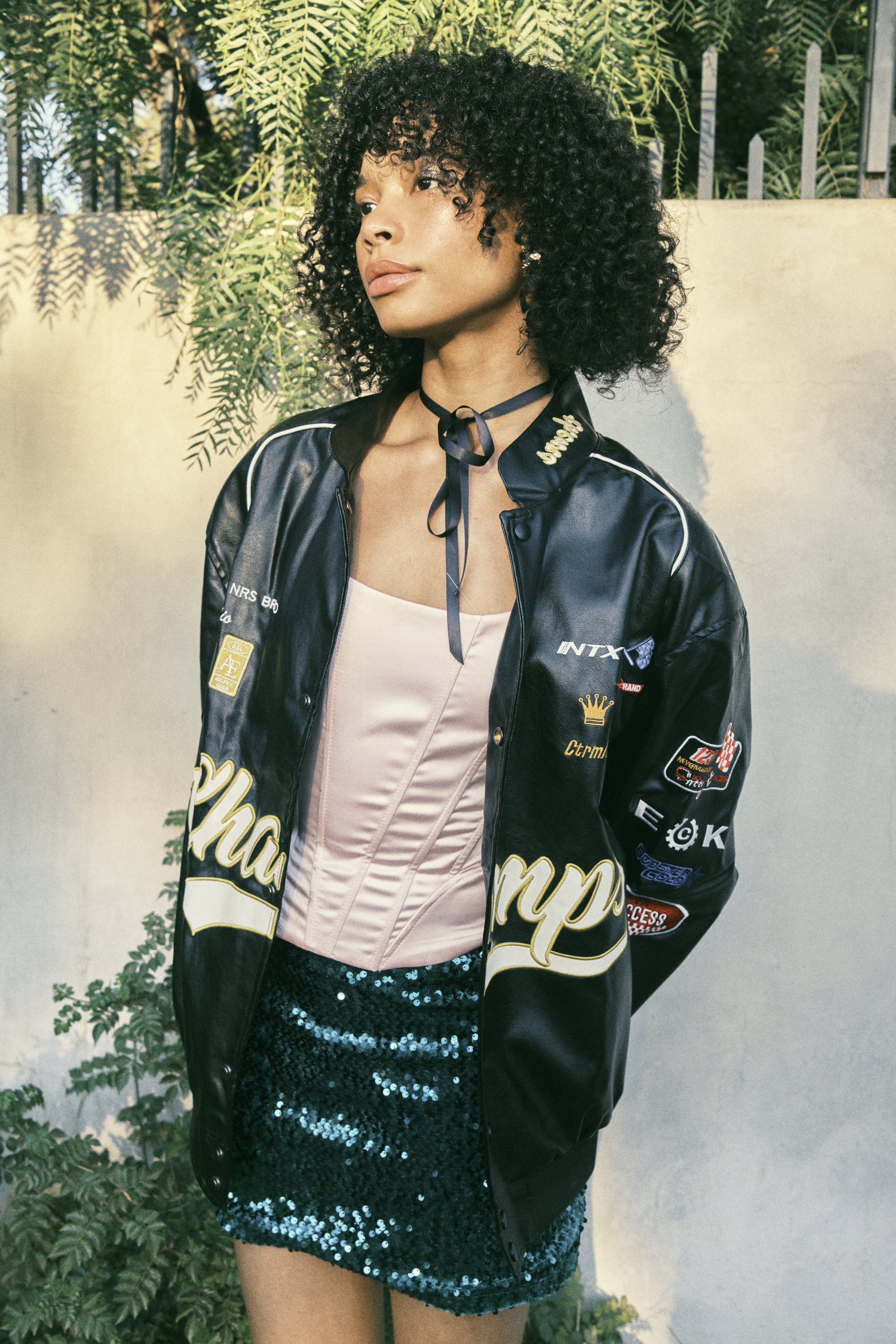 Portrait of a young woman with curly hair wearing a racing jacket and sequin skirt, posing outdoors against a light-colored wall with greenery.