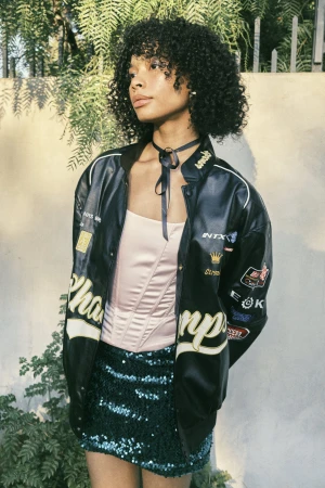 Portrait of a young woman with curly hair wearing a racing jacket and sequin skirt, posing outdoors against a light-colored wall with greenery.