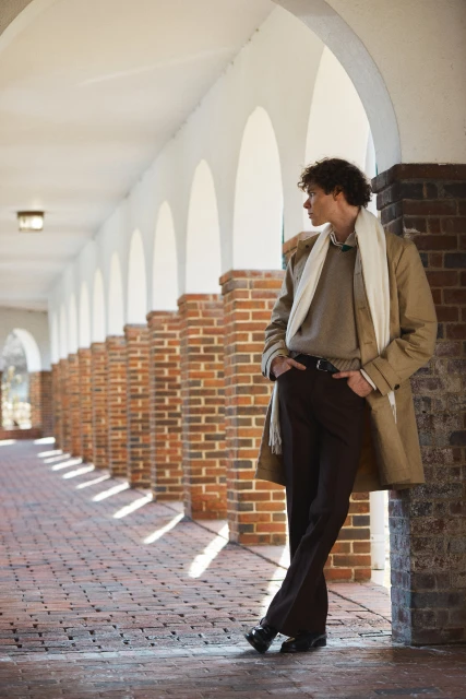Male model leans against a brick column in an arched walkway, wearing a coat, scarf, sweater, and brown slacks, with hands in his pockets.
