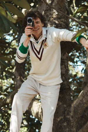 Man with curly brown hair in a white rugby shirt holding a vintage movie camera while climbing in a tree.