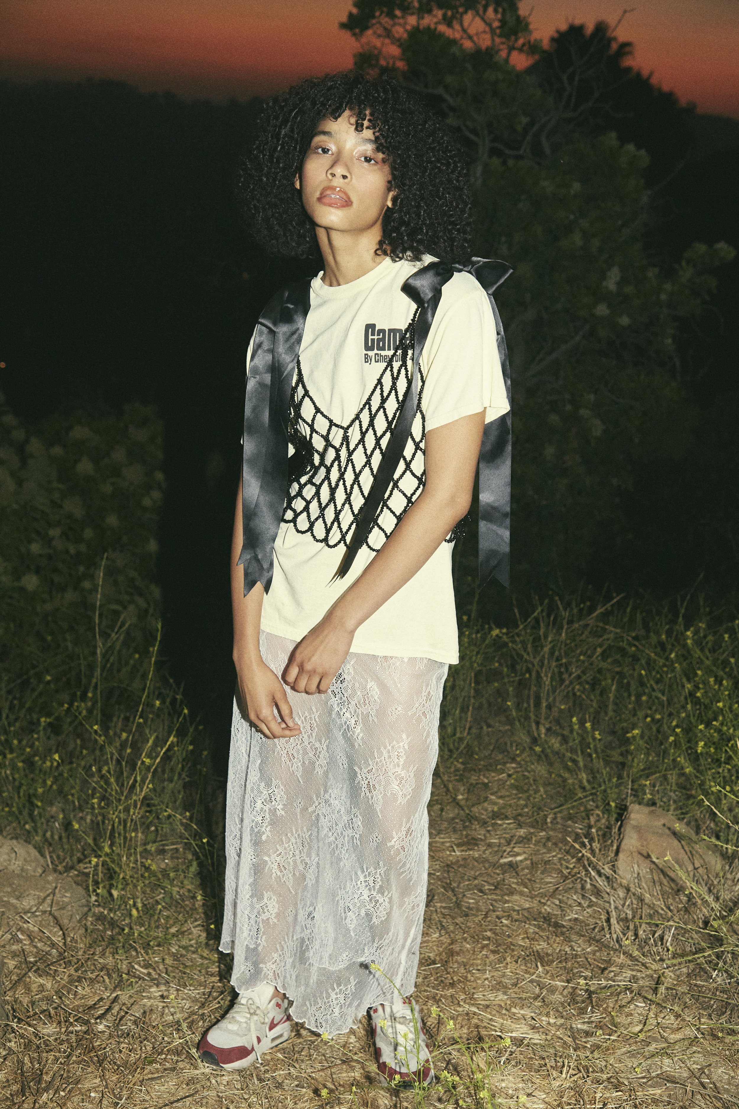 Woman poses outdoors in a pale-yellow T-shirt, black netting vest, sheer lace skirt, and sneakers, against a sunset-lit horizon backdrop.