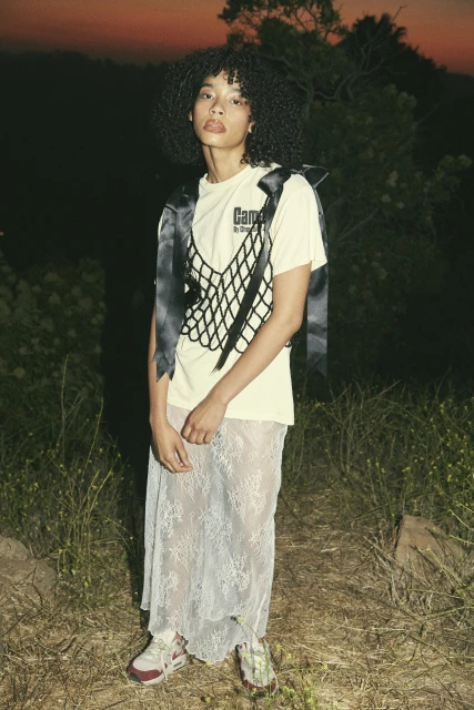 Woman poses outdoors in a pale-yellow T-shirt, black netting vest, sheer lace skirt, and sneakers, against a sunset-lit horizon backdrop.