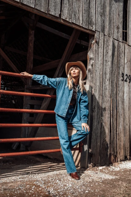 Woman in denim jacket and cowboy hat poses near a weathered barn, showcasing vintage fashion in natural light. Farm setting.