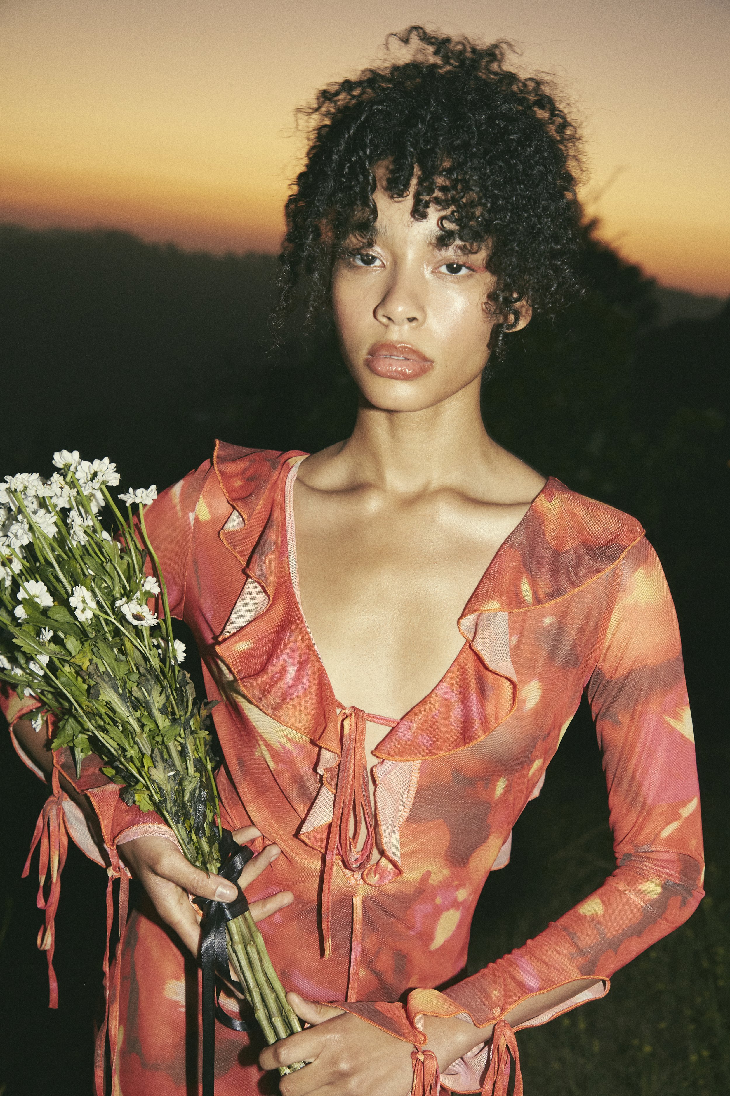 Young woman in orange floral dress holds a bouquet of daisies against a dusky sky at dusk. Fashion shot with curly hair.