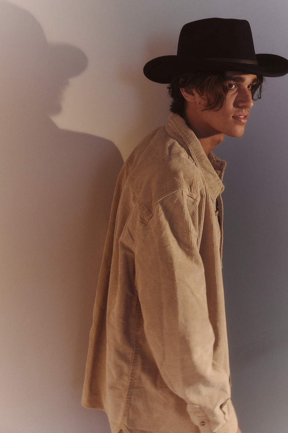 A young man wearing a black hat and beige corduroy shirt looking sideways against a textured wall with shadow.