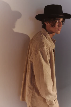 A young man wearing a black hat and beige corduroy shirt looking sideways against a textured wall with shadow.