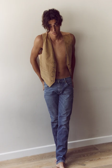 Young adult man in blue jeans and a suede vest stands against a wall in a fashion pose. The scene is lit by natural light.
