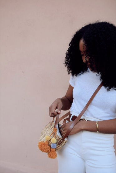 Close-up of a Black woman wearing a white shirt and pants, opening a woven handbag with a leather strap against a neutral wall.