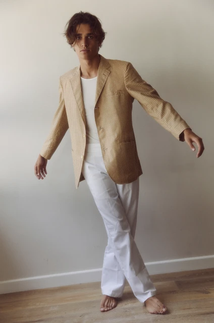 Man in yellow patterned blazer and white pants strikes a pose against a neutral wall, illuminated by natural light on a wooden floor.