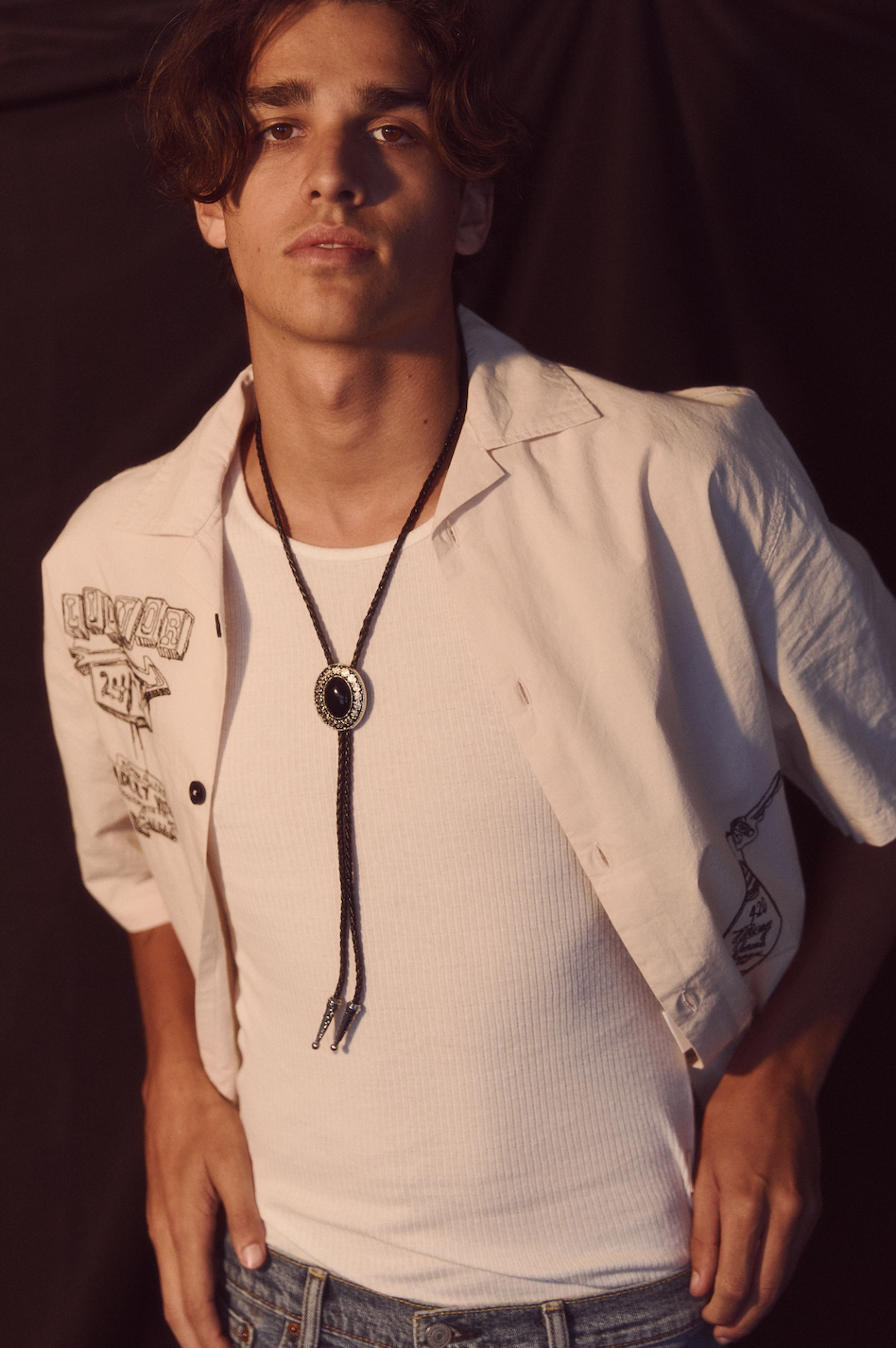 Portrait of a young adult male with dark hair wearing a white shirt and bolo tie against a dark background.