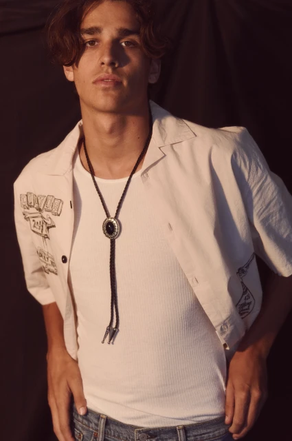 Portrait of a young adult male with dark hair wearing a white shirt and bolo tie against a dark background.