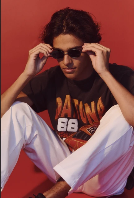 Young adult in sunglasses and a graphic tee poses against a solid red backdrop, showcasing a relaxed yet stylish casual fashion statement.