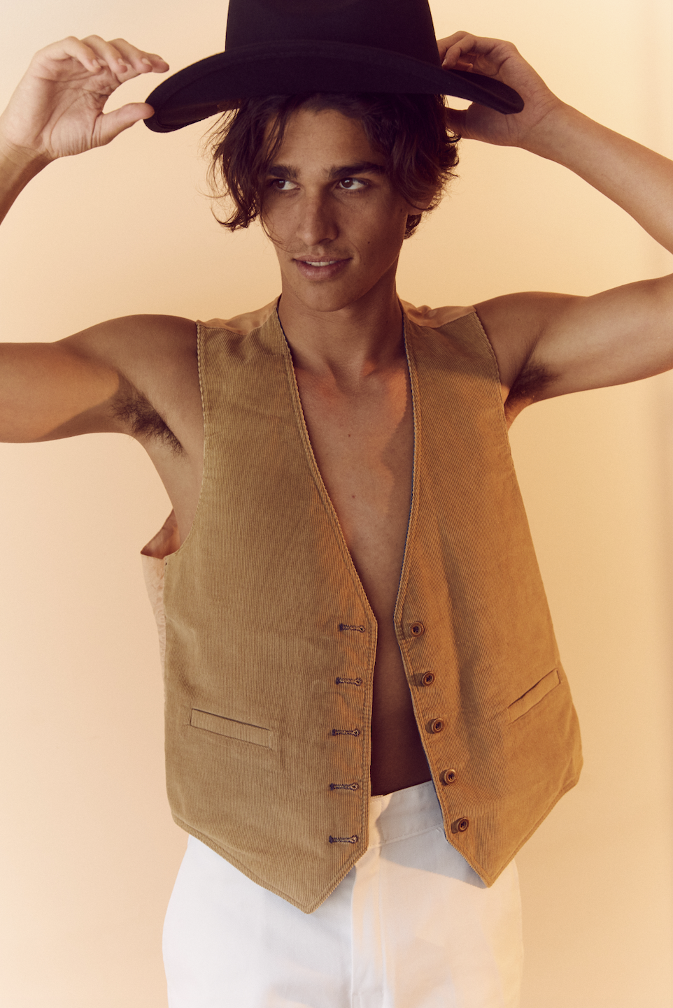 A young adult male with a dark hat and tan vest poses in front of a warm-toned backdrop, looking slightly off-camera.