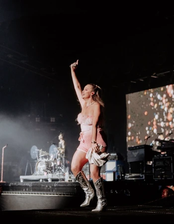 A blonde female performer stands on stage in a pink outfit and cowboy boots, holding a cloth, with a band visible in the background.