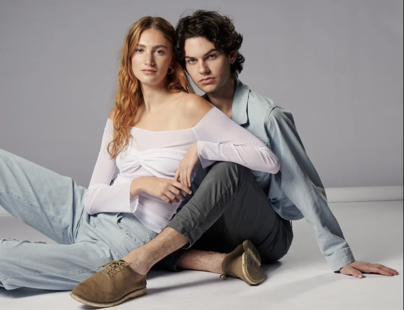 A woman and a man sit close together on a white floor against a grey background. The woman wears a white top and striped pants.