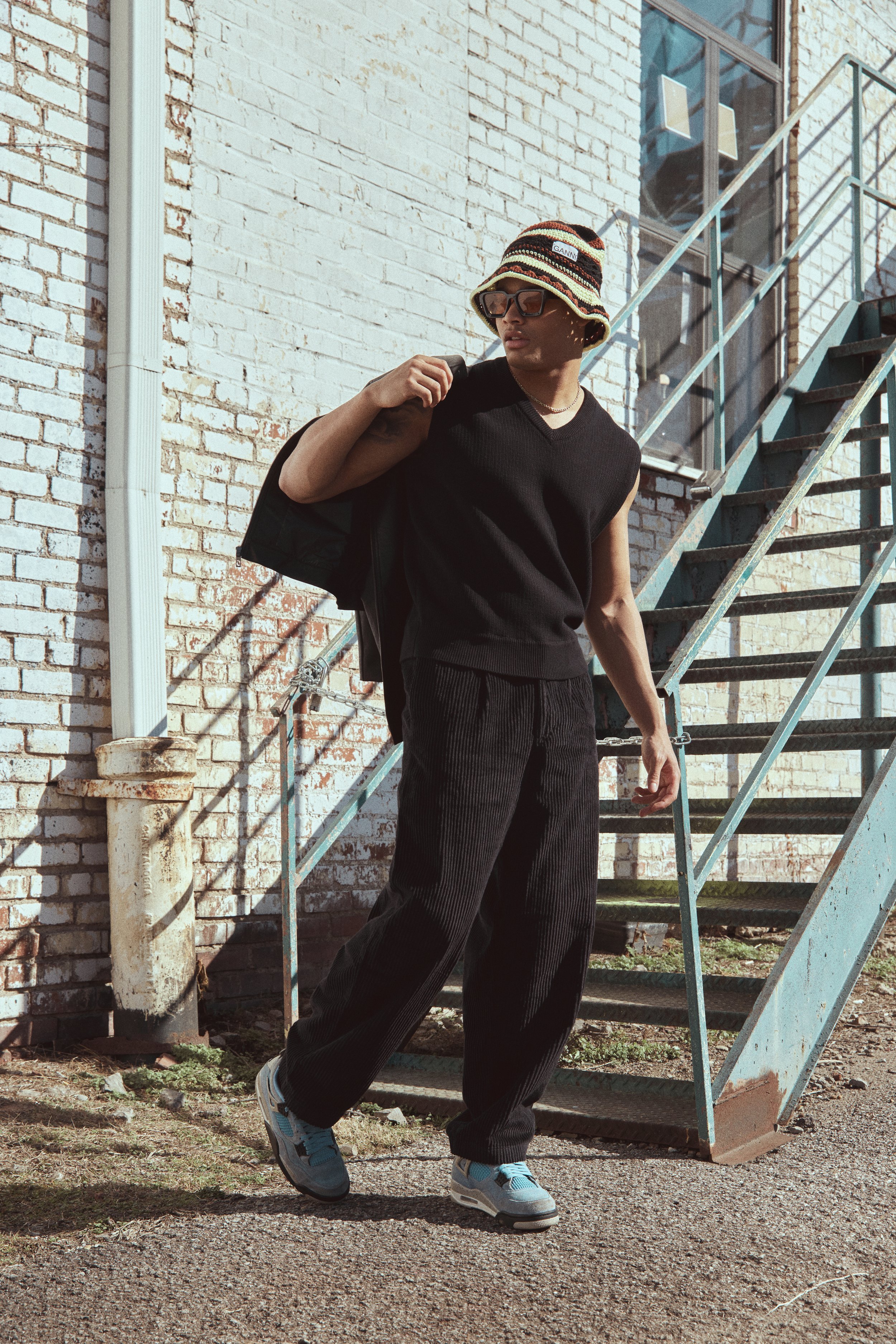 Young man in casual fashion poses near a brick building with black vest, bucket hat, and sneakers, exuding casual style.